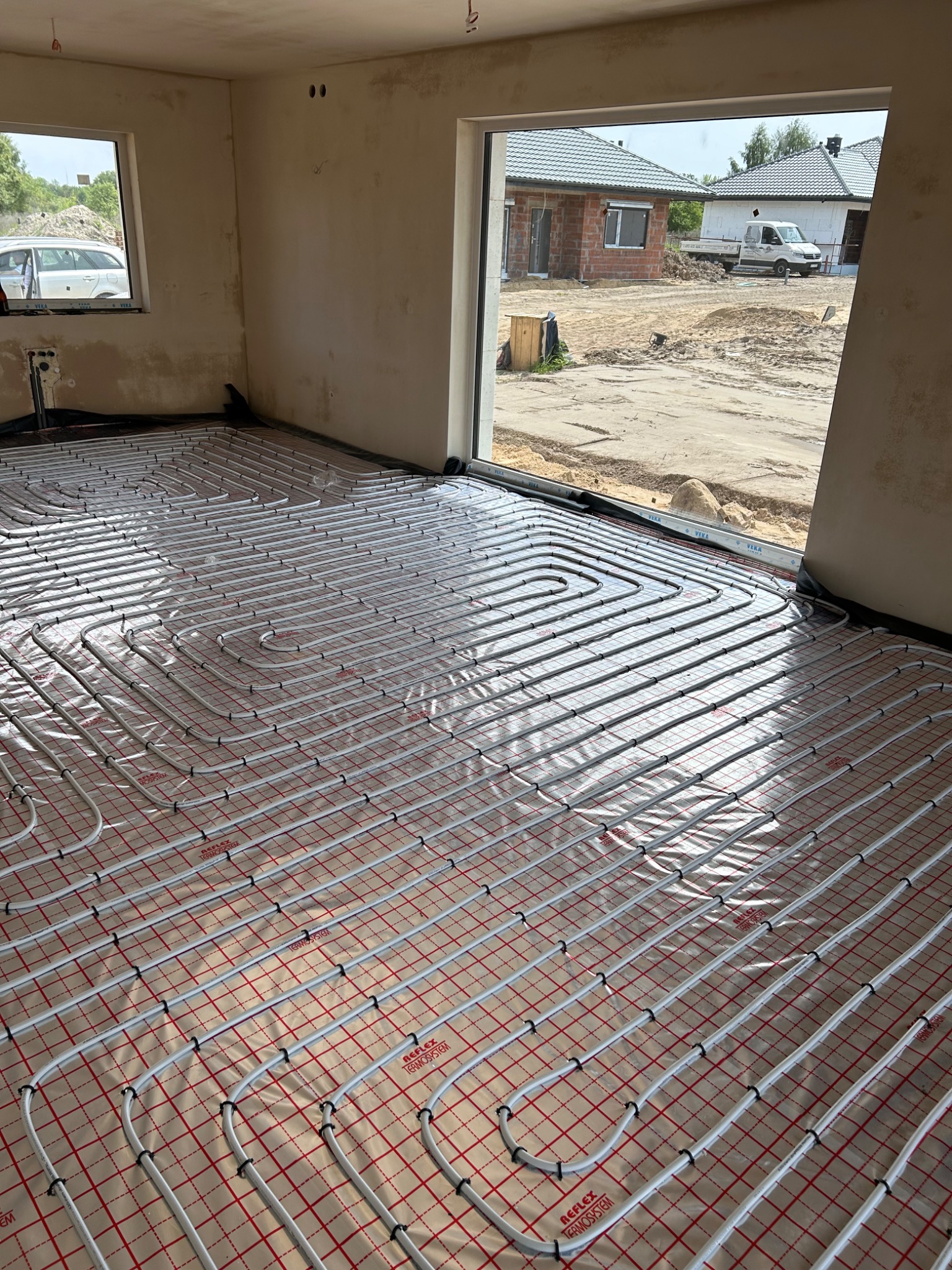 Białe rury grzewcze ułożone w pętle na folii izolacyjnej z czerwoną siatką, widoczne w surowym wnętrzu z oknami wychodzącymi na plac budowy.
