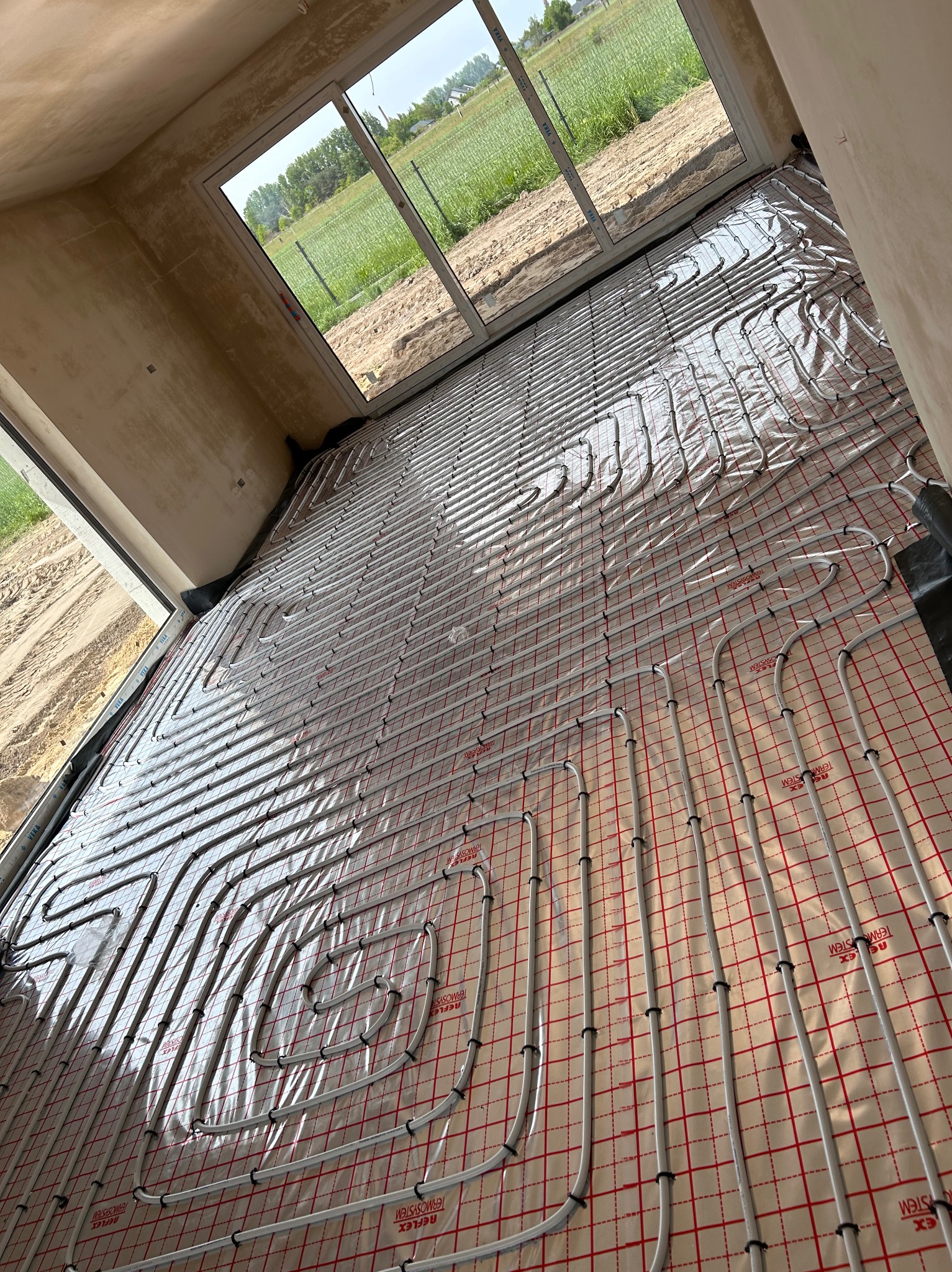 Białe rury ogrzewania podłogowego ułożone na folii z czerwoną siatką w surowym wnętrzu z dużym oknem wychodzącym na zielone pole.