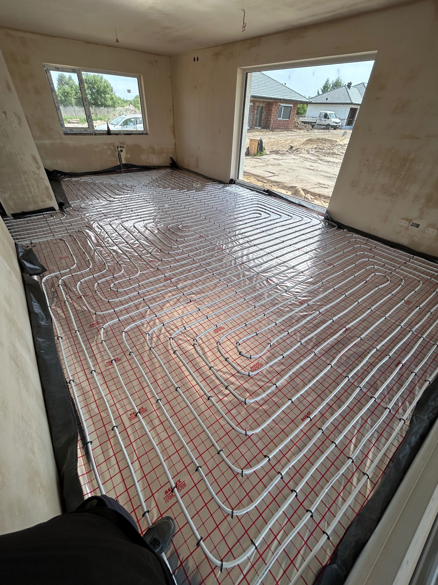 Białe rurki ogrzewania podłogowego ułożone na czerwonej siatce w surowym wnętrzu, widok na okno z budynkiem w tle, czarna folia na brzegach podłogi.