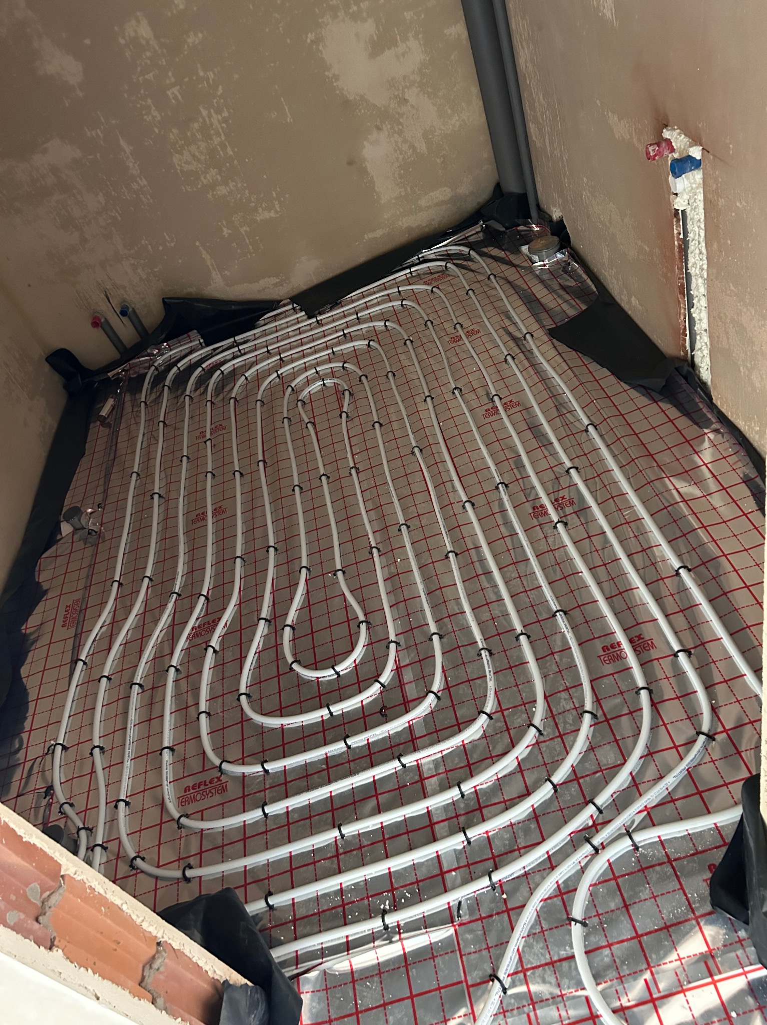 Białe rury ogrzewania podłogowego ułożone na folii z czerwoną siatką, widoczne mocowania rur i fragment ściany w surowym stanie.