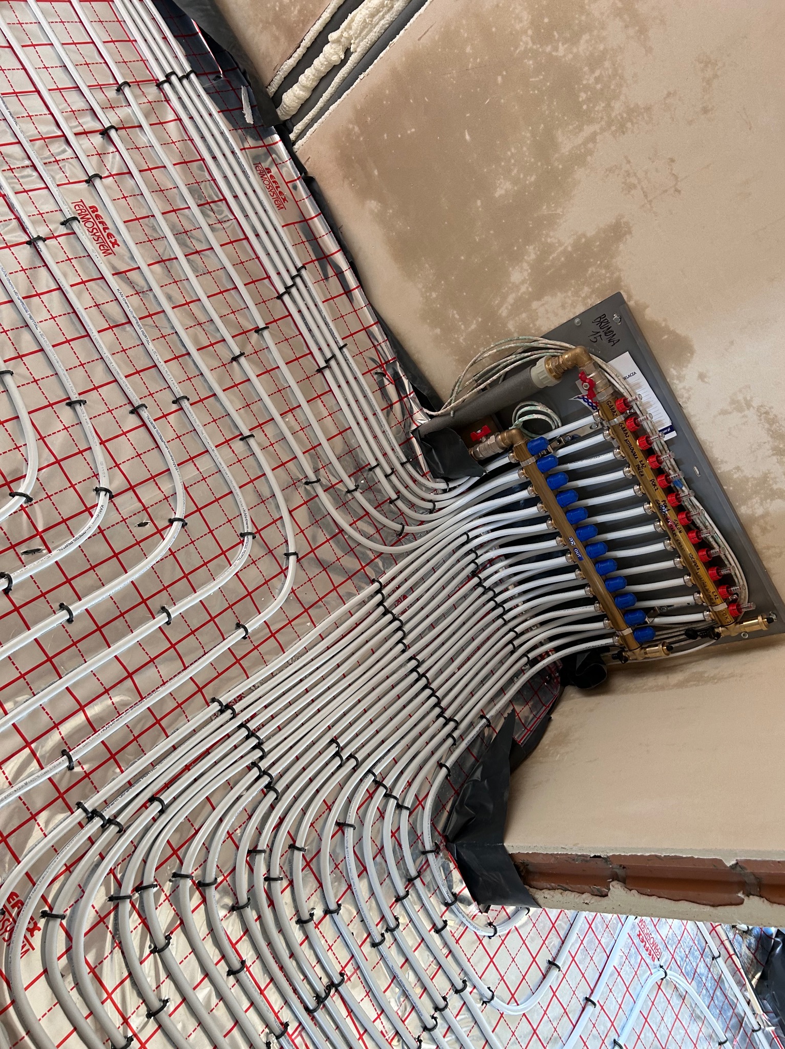 Rozłożona instalacja ogrzewania podłogowego z widocznym rozdzielaczem, rurami PEX przymocowanymi do folii refleksyjnej z nadrukiem w czerwoną kratkę. Ściana w stanie surowym w tle.