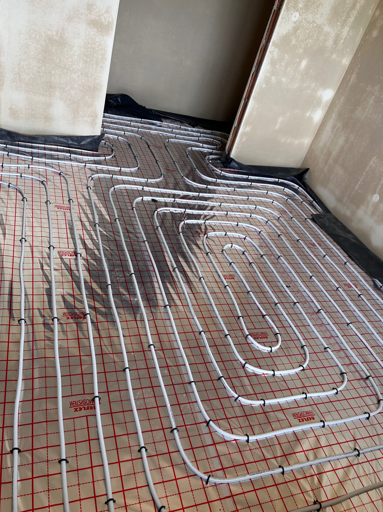 Białe rury ogrzewania podłogowego ułożone na folii z czerwoną kratką, przymocowane czarnymi klipsami, widoczne przygotowania do zalania wylewką.