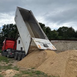 FIRMA TRANSPORTOWA IWONA CZOCHARA - Czerwona ciężarówka z podniesioną naczepą wysypuje piasek na placu budowy; widoczne drzewa i budynki w tle, pochmurne niebo.