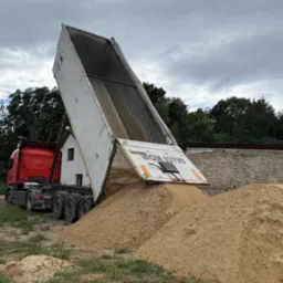 Czerwona ciężarówka z podniesioną naczepą wysypuje piasek na placu budowy; widoczne drzewa i budynki w tle, pochmurne niebo.