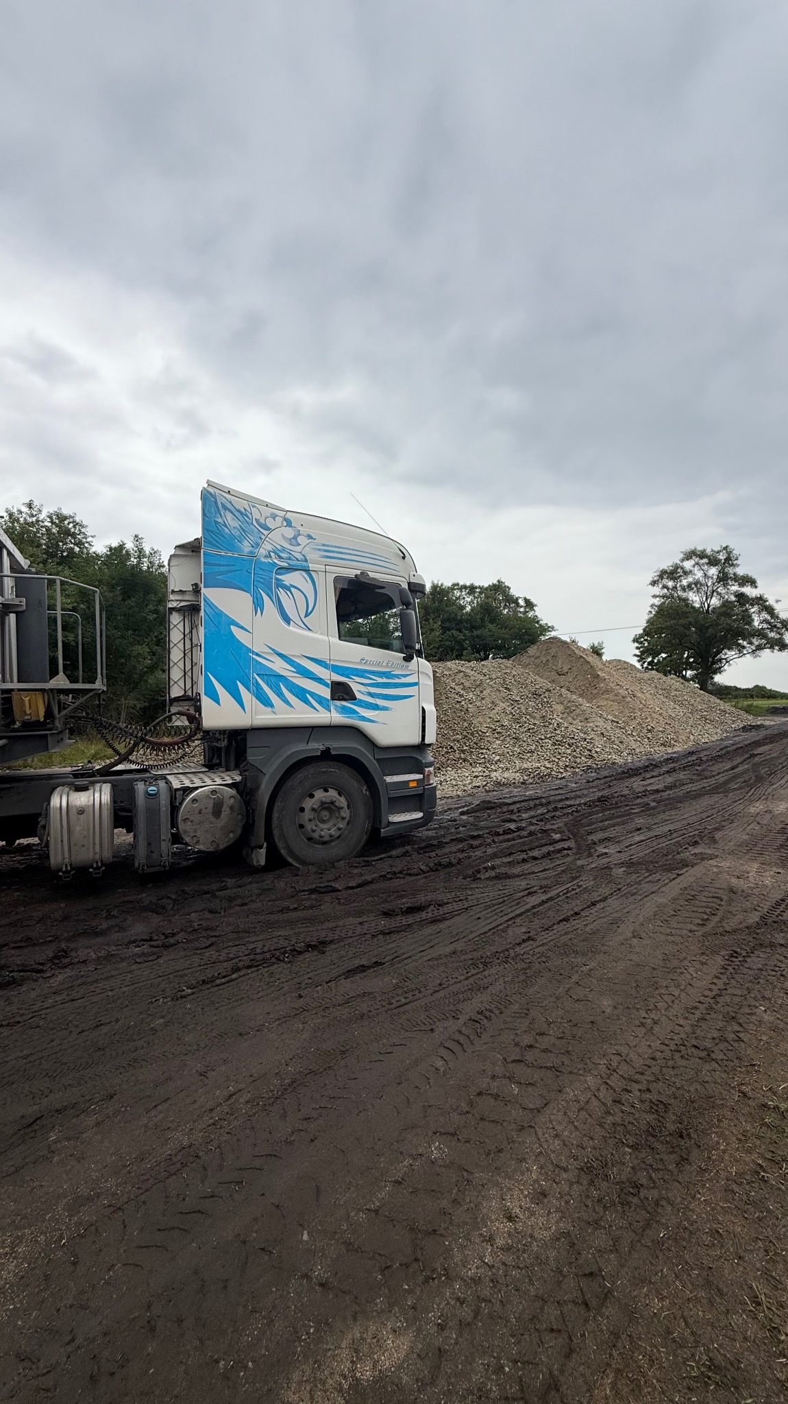 Ciężarówka z niebieskim motywem graficznym na kabinie, stojąca na błotnistej drodze obok hałdy żwiru, w tle drzewa i pochmurne niebo.