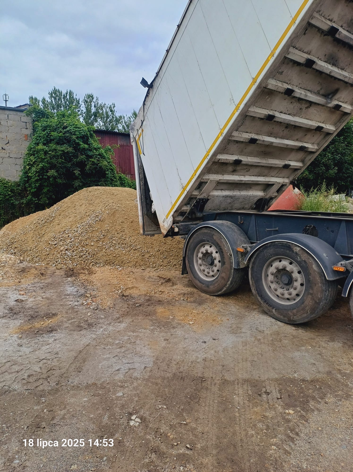 Wyładowany piasek z białej naczepy ciężarówki na placu budowy. Widoczne opony ciężarówki, w tle zieleń i zabudowania. Ujęcie z dołu.