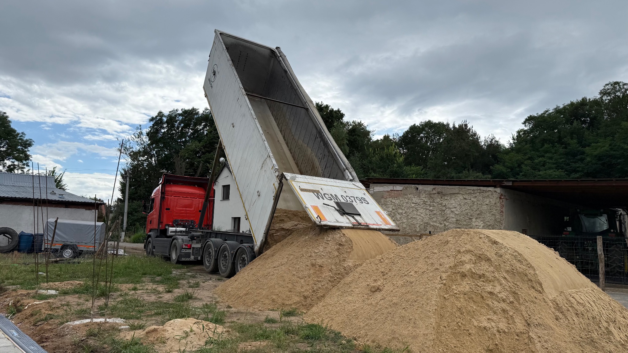 Czerwona ciężarówka z podniesioną naczepą wysypuje piasek na placu budowy; widoczne drzewa i budynki w tle, pochmurne niebo.