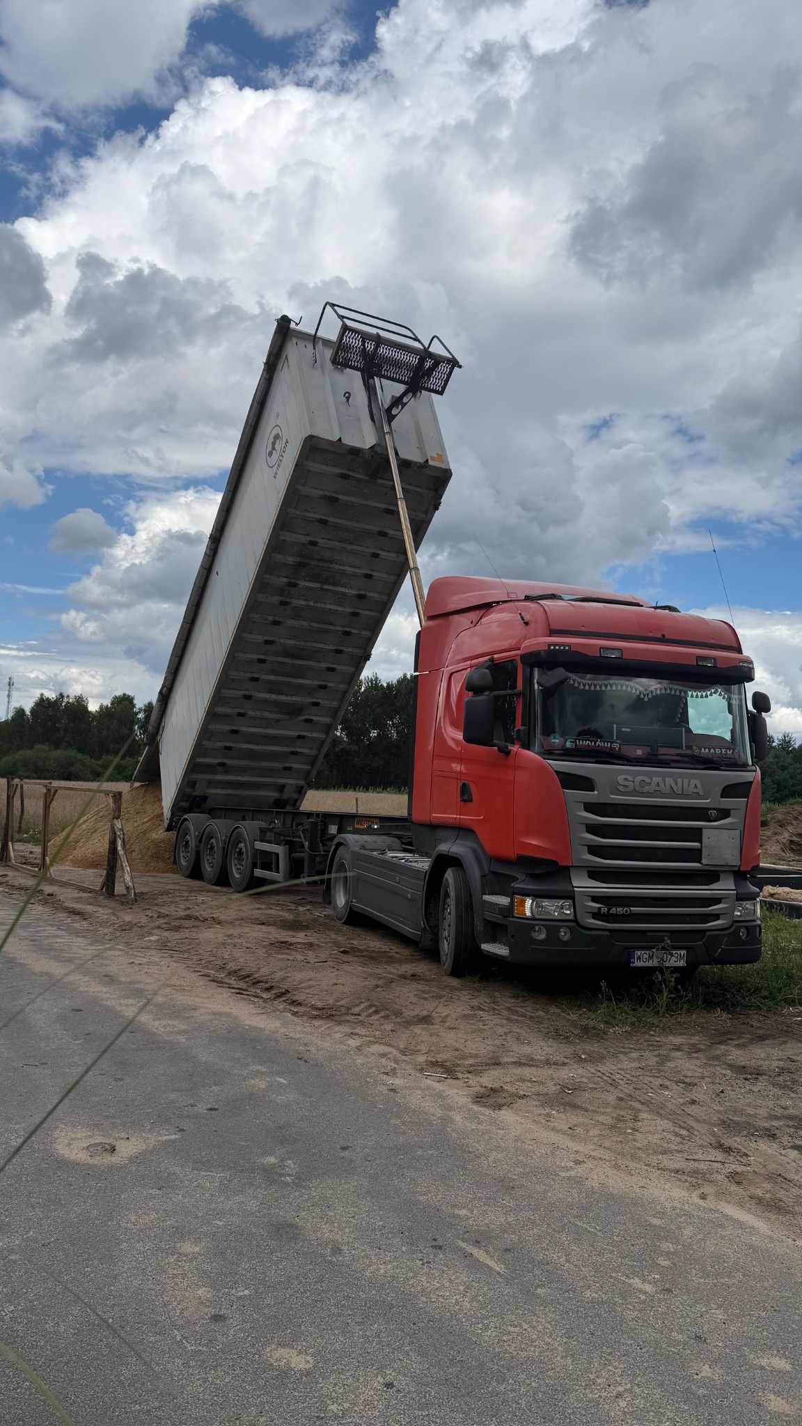 Czerwona ciężarówka Scania R450 z podniesioną naczepą wyładowuje piasek na tle pochmurnego nieba i zieleni. Widoczny fragment drogi i ogrodzenie.