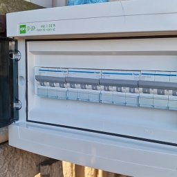 Damian Dąbrowski - Otwarta skrzynka elektryczna z bezpiecznikami Hager, zamontowana na betonowej ścianie z widocznym drewnianym elementem mocującym, ukazująca wnętrze z kilkoma wyłącznikami różnicowoprądowymi.