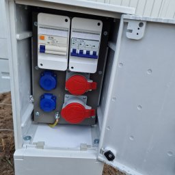 Damian Dąbrowski - Otwarta, biała skrzynka elektryczna na zewnątrz z dwoma wyłącznikami LC TEC i czterema gniazdami (dwa niebieskie, dwa czerwone) zamontowanymi na szarej płycie, widoczne przewody uziemiające.