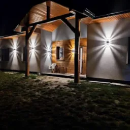 Nocne ujęcie elewacji domu z zamontowanymi lampami zewnętrznymi, drewnianą konstrukcją zadaszenia nad wejściem i oświetlonym wejściem, widoczne krzesło i stolik.