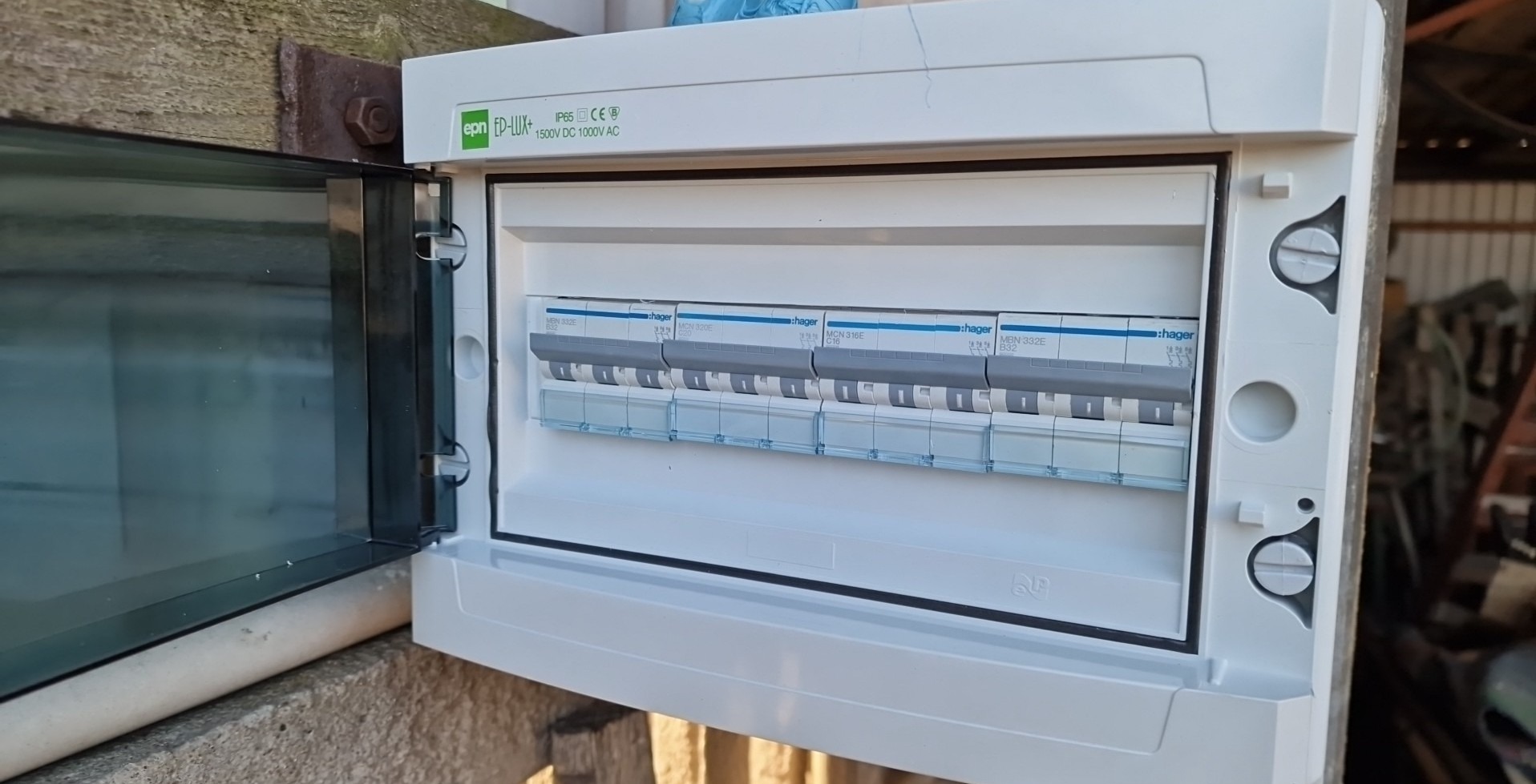 Otwarta skrzynka elektryczna z bezpiecznikami Hager, zamontowana na betonowej ścianie z widocznym drewnianym elementem mocującym, ukazująca wnętrze z kilkoma wyłącznikami różnicowoprądowymi.