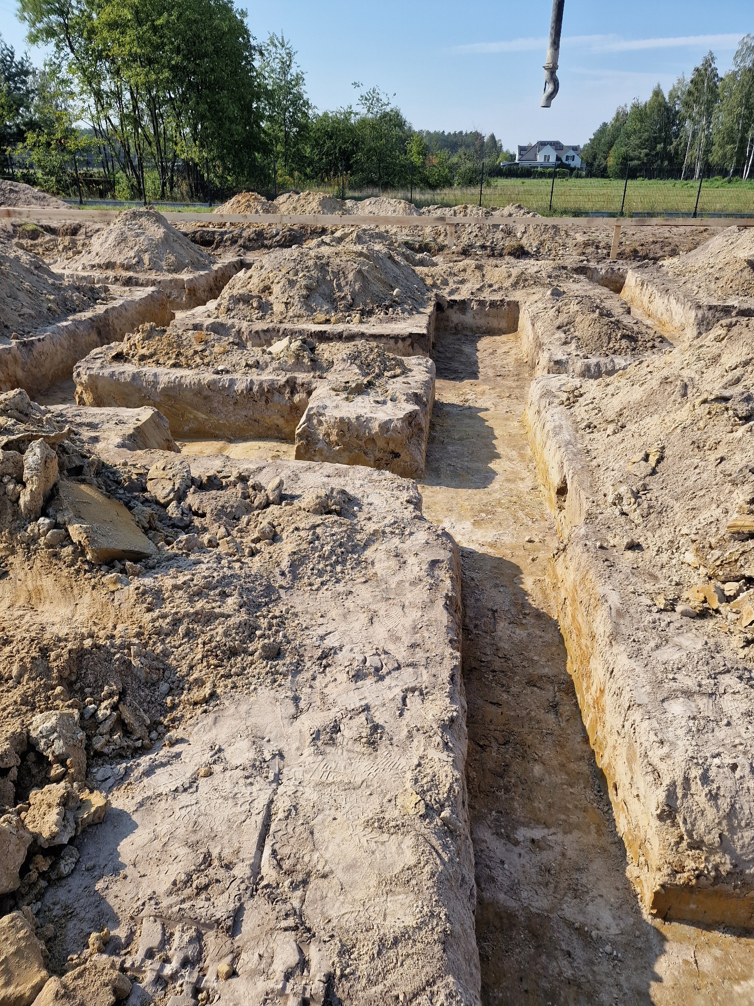 Wykopane rowy pod fundamenty z widocznymi śladami opon i stertami ziemi, w tle dom jednorodzinny i drzewa pod błękitnym niebem.