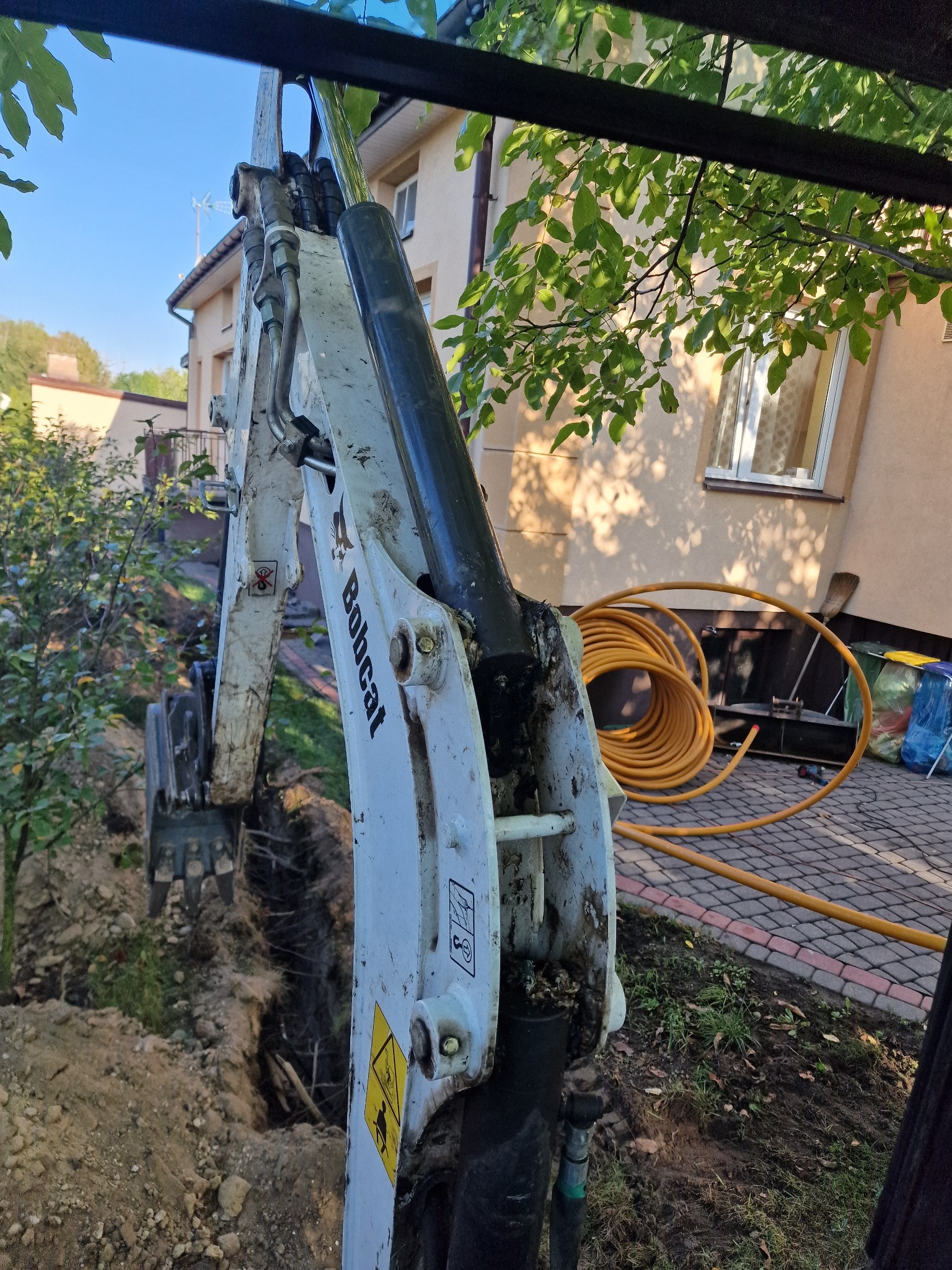 Mini koparka Bobcat przy wykopie w ziemi, w tle dom jednorodzinny i zwinięty żółty wąż.