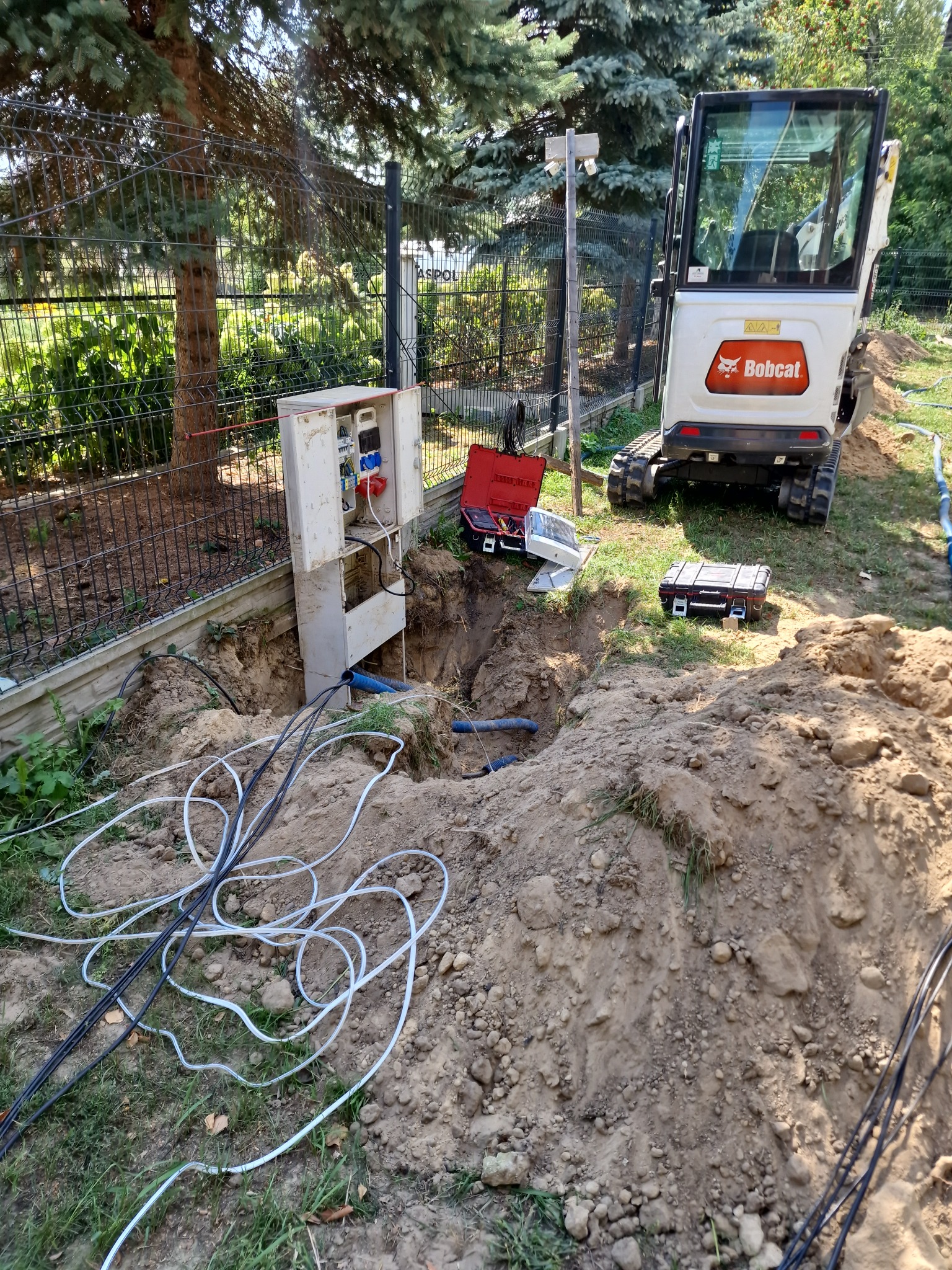 Szafka elektryczna z otwartymi drzwiczkami, wystającymi kablami i podłączonymi wtyczkami, umieszczona obok wykopu; w tle minikoparka Bobcat i ogrodzenie z siatki.