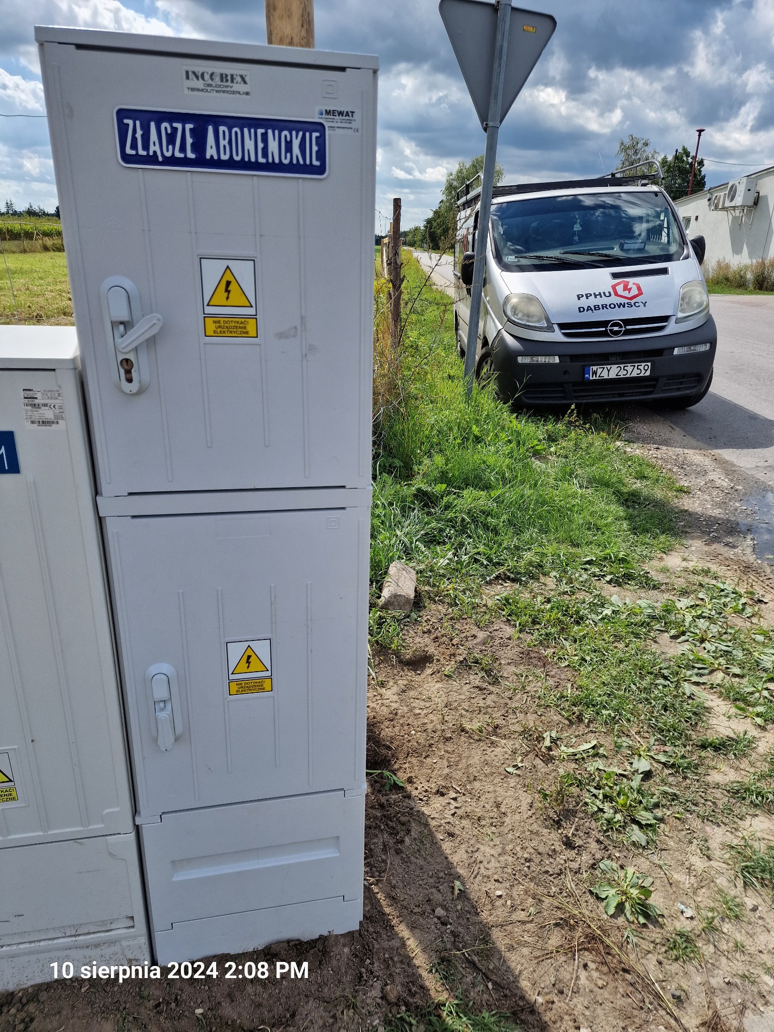 Szara szafa z napisem 'Złącze Abonenckie' i naklejkami ostrzegawczymi, obok zaparkowany srebrny samochód dostawczy z logo firmy elektrycznej na tle pochmurnego nieba.