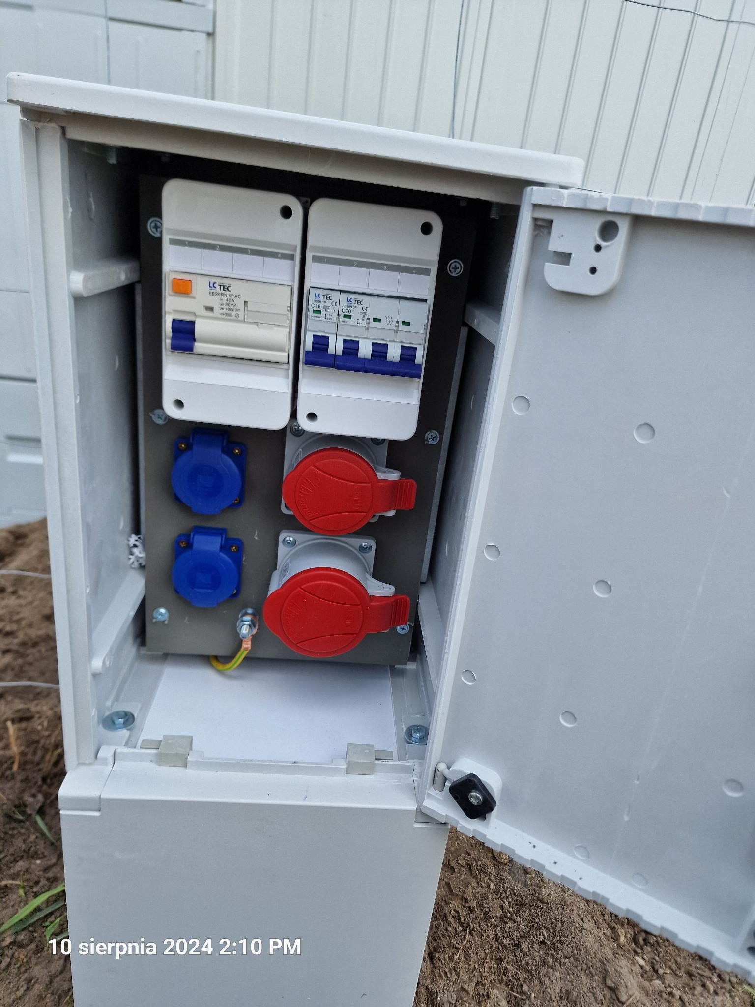 Otwarta, biała skrzynka elektryczna na zewnątrz z dwoma wyłącznikami LC TEC i czterema gniazdami (dwa niebieskie, dwa czerwone) zamontowanymi na szarej płycie, widoczne przewody uziemiające.