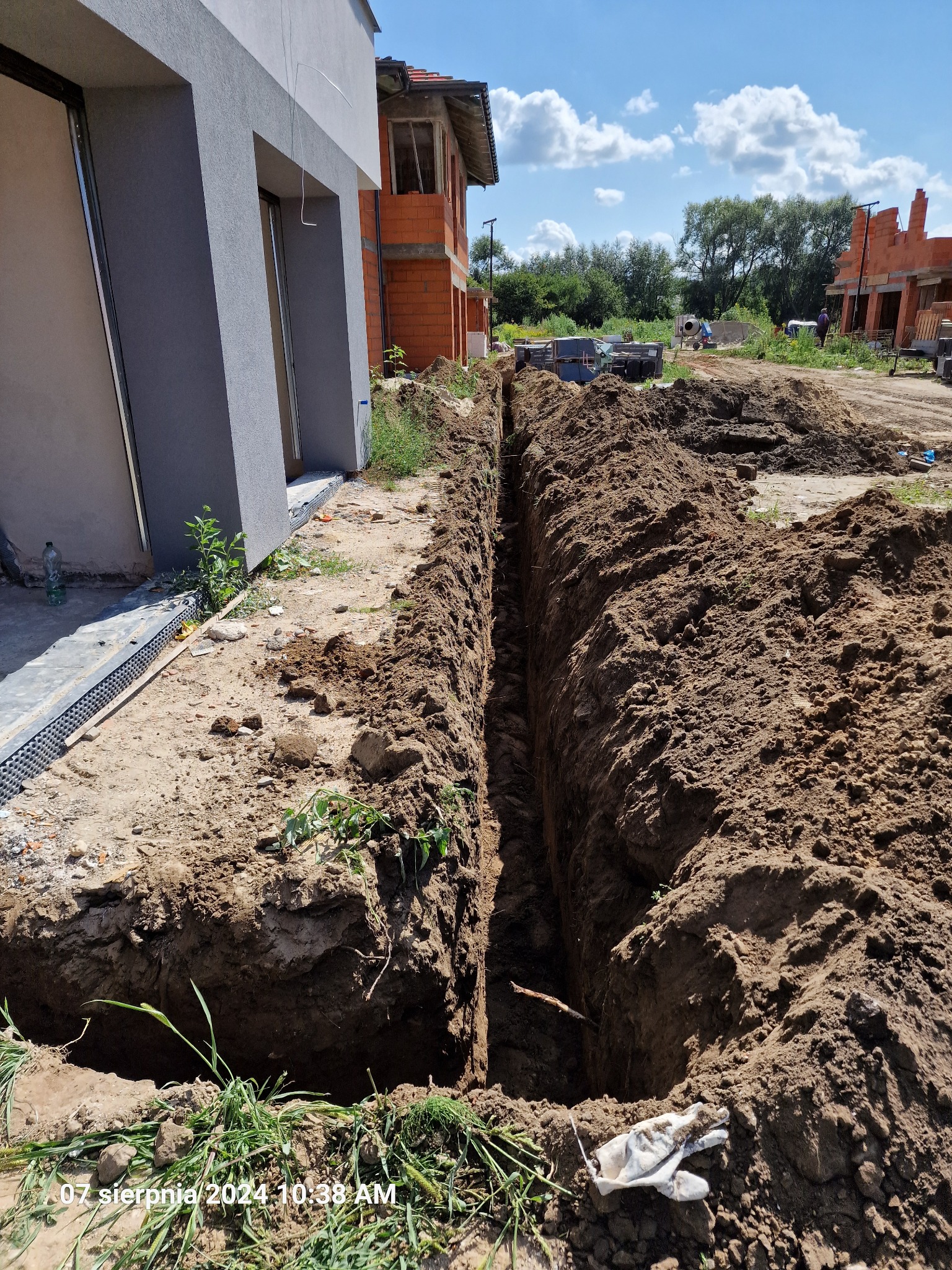 Wąski wykop w ziemi, prawdopodobnie pod instalacje, biegnący wzdłuż fundamentów budynków w stanie surowym otwartym, widoczne ślady prac ziemnych i elementy izolacji fundamentów.