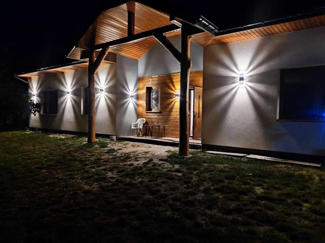 Nocne ujęcie elewacji domu z zamontowanymi lampami zewnętrznymi, drewnianą konstrukcją zadaszenia nad wejściem i oświetlonym wejściem, widoczne krzesło i stolik.