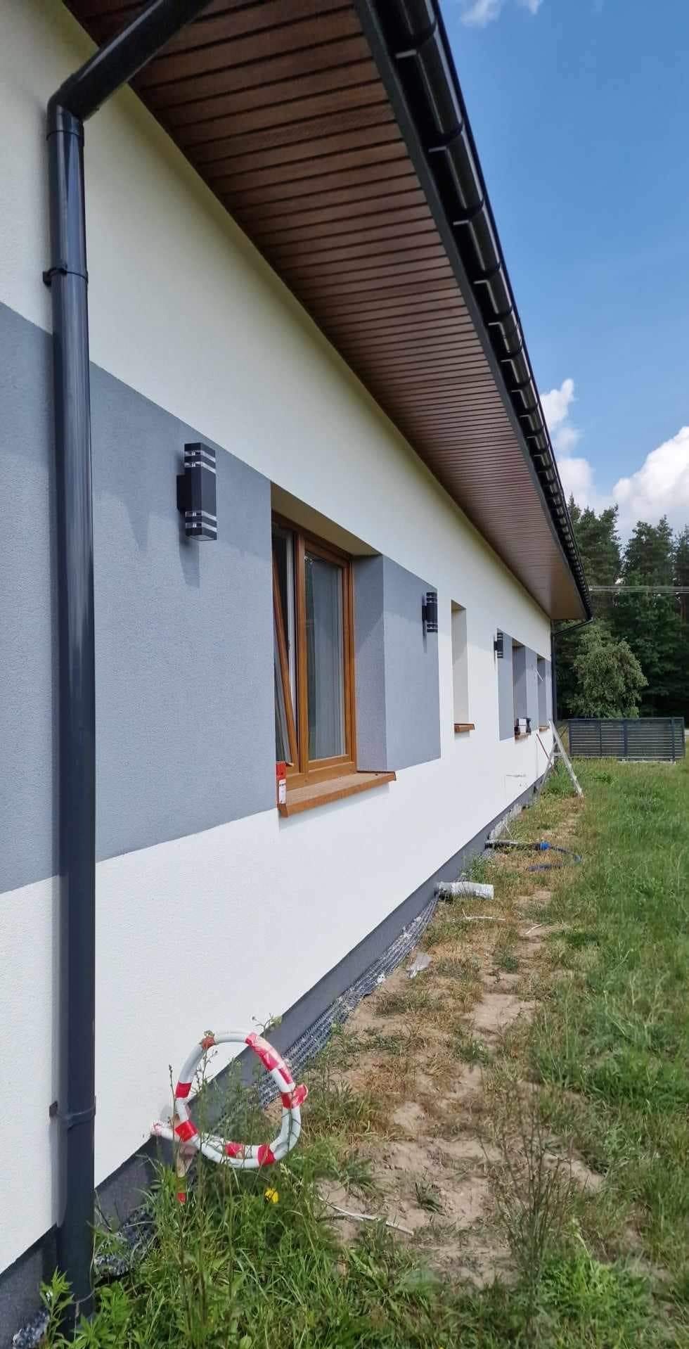 Elewacja domu jednorodzinnego z zamontowanym oświetleniem zewnętrznym, widoczne okna z drewnianą stolarką, orynnowanie i fragment terenu z trawą i elementami zabezpieczającymi.