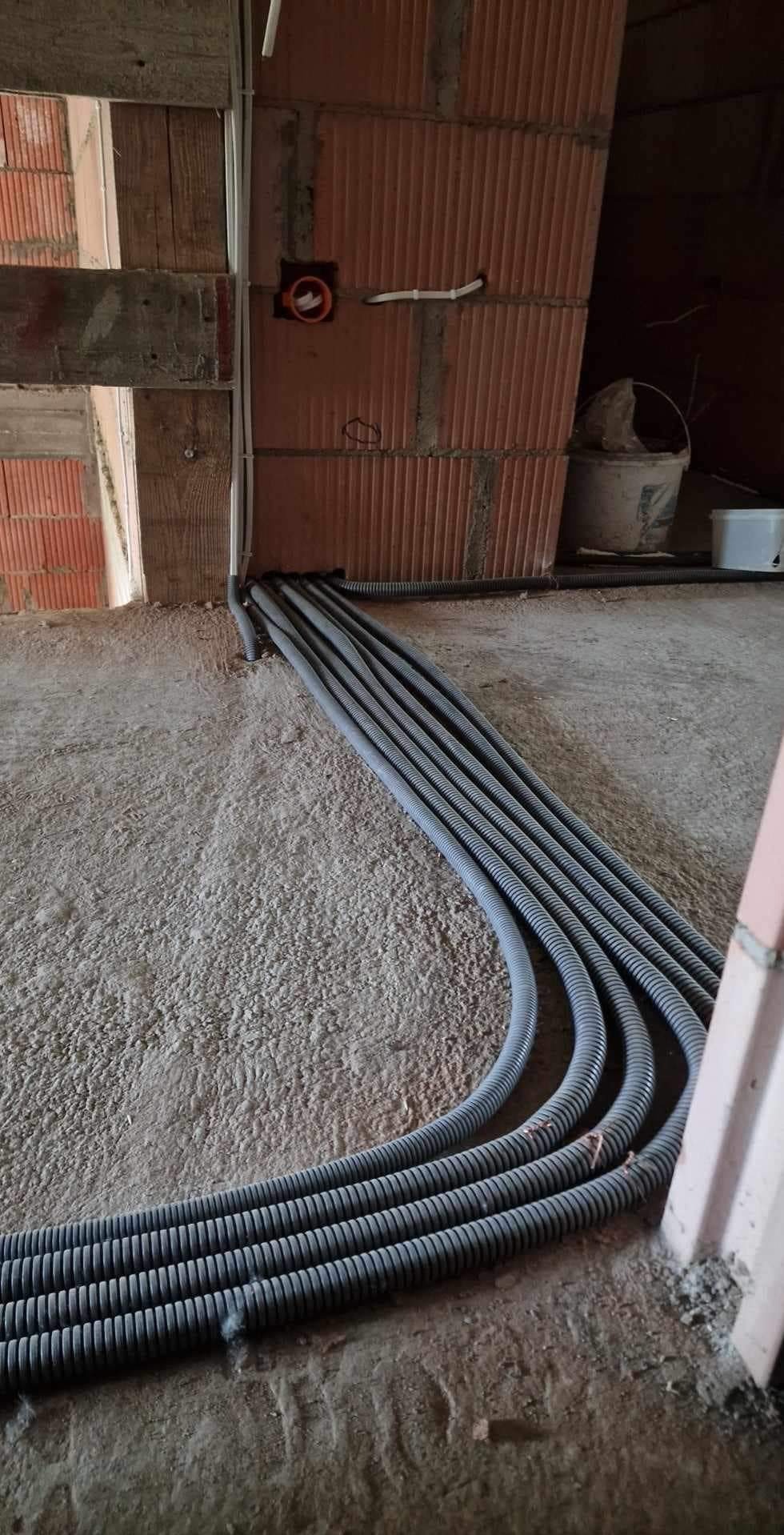 Równe rzędy szarych, karbowanych rur instalacyjnych wychodzących ze ściany z cegły dziurawki w surowym stanie, prowadzone po betonowej podłodze w nowo budowanym budynku.