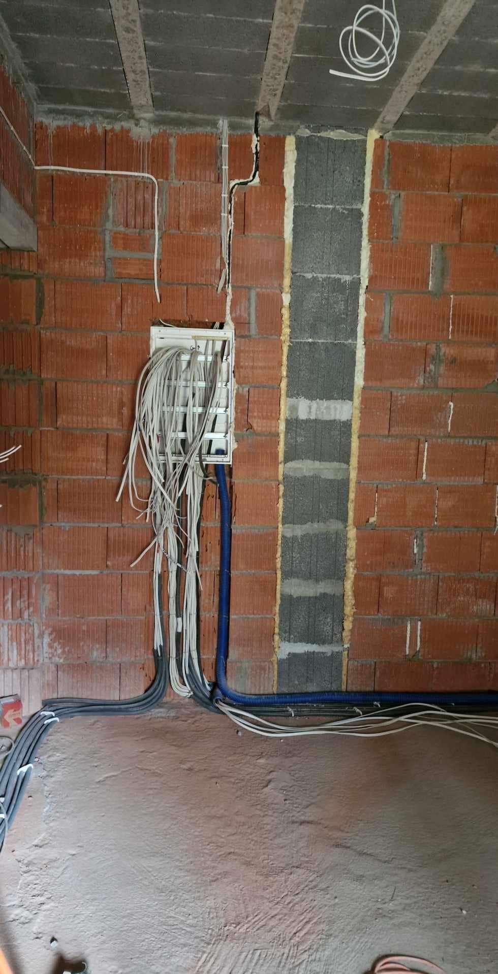 Widok na ścianę z czerwonej cegły z zamontowaną białą skrzynką elektryczną, z której wychodzą liczne przewody; niebieskie rury ochronne prowadzą wzdłuż ściany i podłogi w surowym stanie.