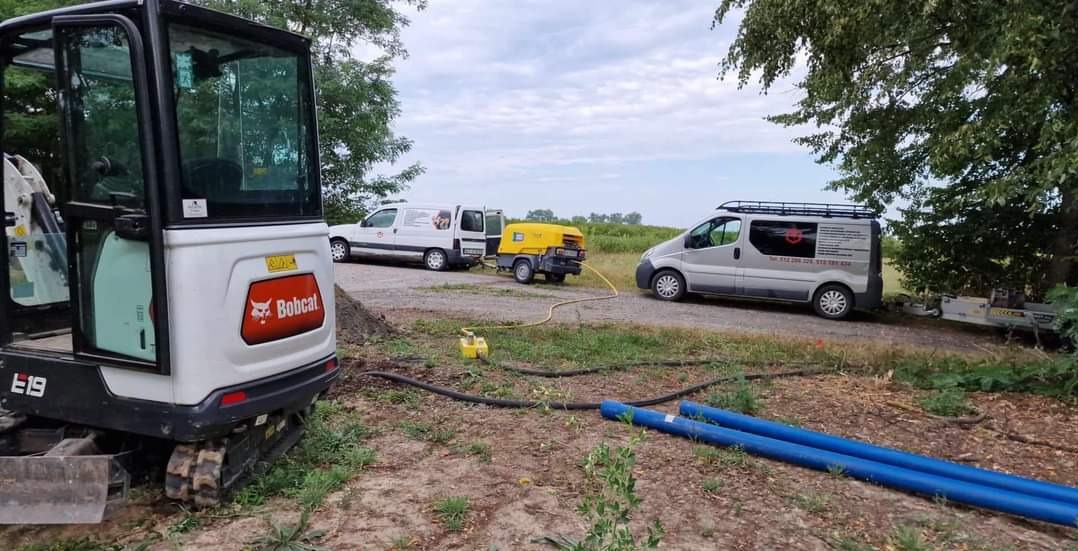 Bobcat E19 mini excavator with open door, compressor connected to a white van, silver van with company logo and contact information, blue pipes on the ground, suggesting excavation or utility...