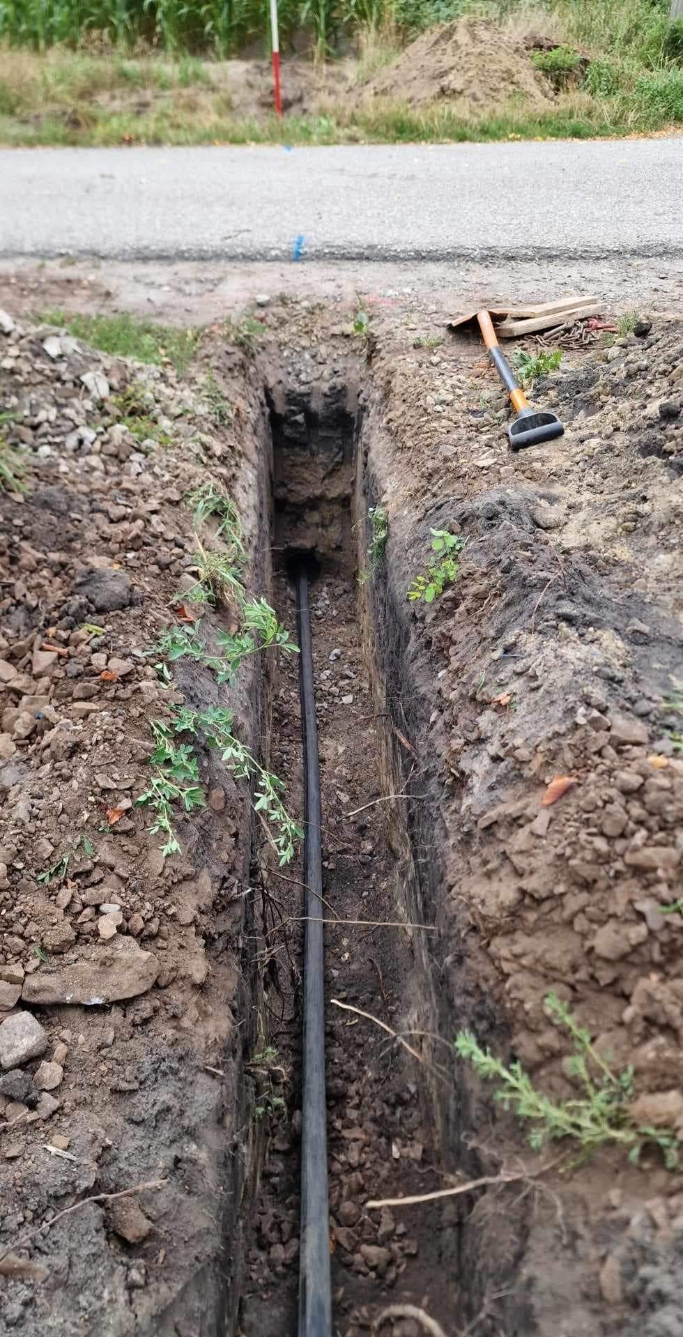 Wąski wykop w ziemi z ułożonym czarnym kablem, widoczne narzędzia i fragment drogi w tle.