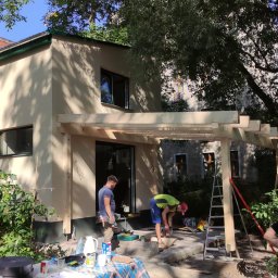 Bau4u - Budowa drewnianej pergoli przy jasnym, otynkowanym domu jednorodzinnym; trzech mężczyzn pracuje, używając wiertarki i drabiny, na zewnątrz, w słoneczny dzień.