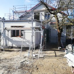 Bau4u - Dom jednorodzinny w trakcie ocieplania elewacji, widoczne rusztowania, fragment drzewa i teren budowy z gruzem.