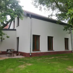 Świeżo otynkowany dom z ceglaną podmurówką, drewnianymi oknami i metalowymi rynnami, widok z zewnątrz na trawnik i drzewa.