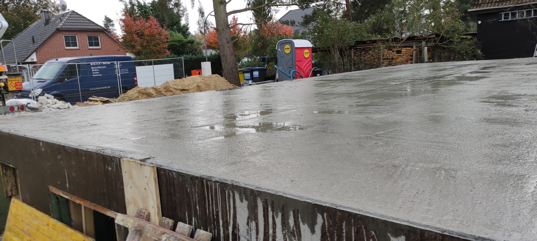 Świeżo wylana, mokra płyta betonowa fundamentowa z widocznym szalunkiem, w tle zaparkowany niebieski bus i przenośna toaleta.