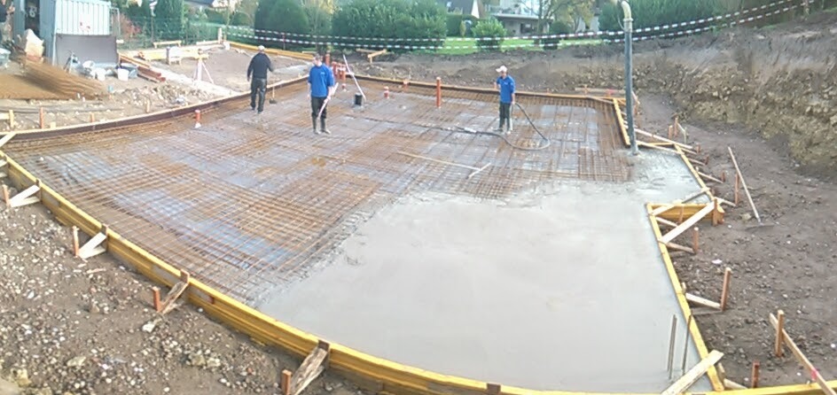 Wylewanie betonu na przygotowane zbrojenie fundamentu pod budowę domu, widoczni pracownicy z narzędziami i szalunki.