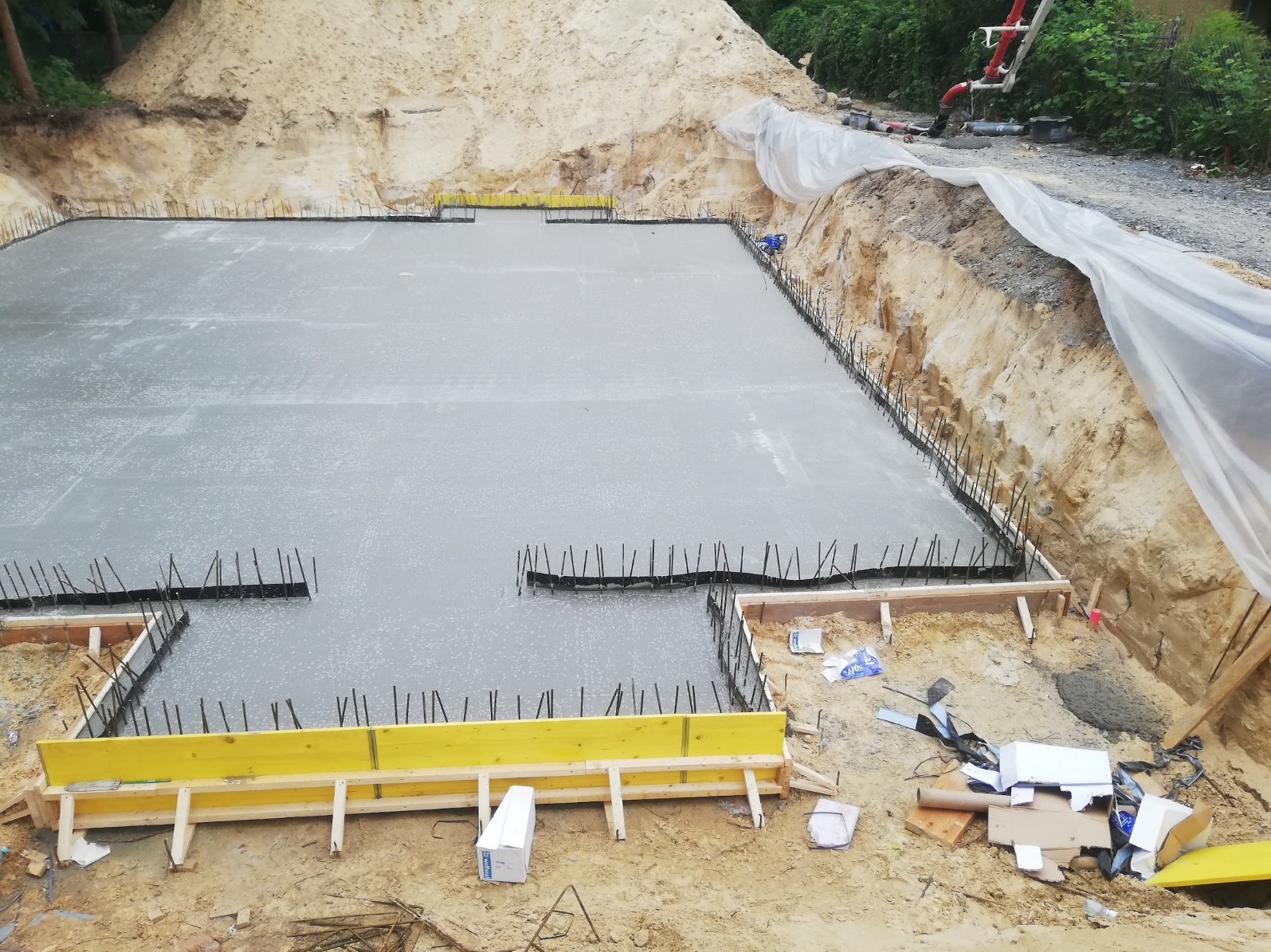 Wylany betonowy fundament z wystającymi prętami zbrojeniowymi, otoczony drewnianym szalunkiem z żółtymi elementami wzmacniającymi, widok z góry na wykop.