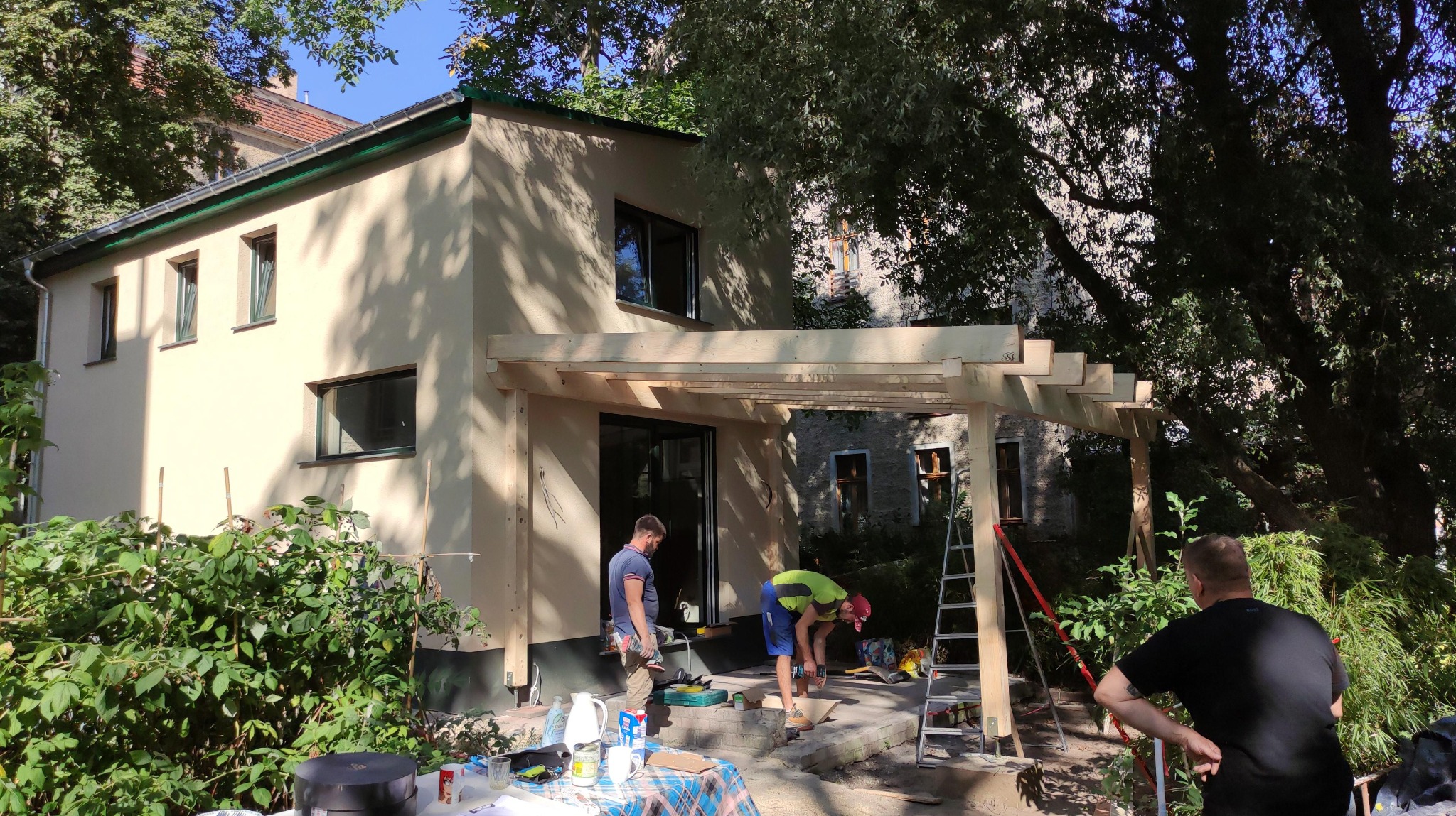 Budowa drewnianej pergoli przy jasnym, otynkowanym domu jednorodzinnym; trzech mężczyzn pracuje, używając wiertarki i drabiny, na zewnątrz, w słoneczny dzień.