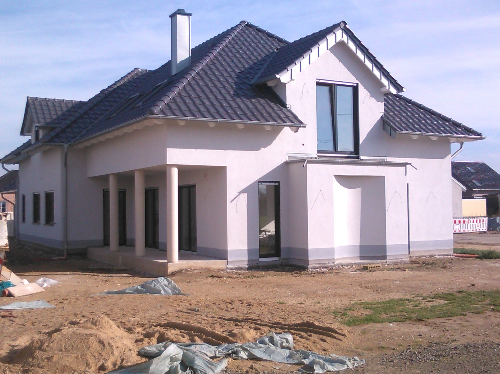 Surowy stan budowy domu jednorodzinnego z ciemną dachówką, jasną elewacją i kolumnami przy wejściu, widoczne ślady prac ziemnych i materiały budowlane na działce.