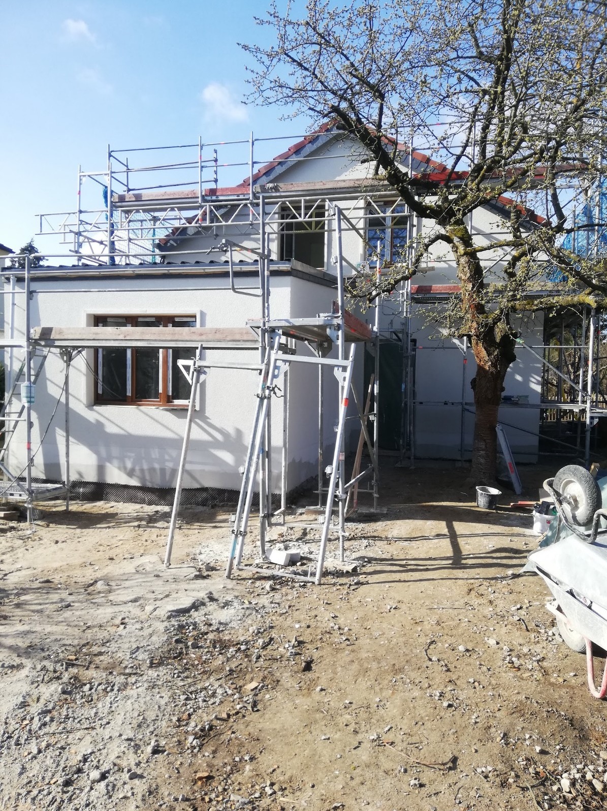 Dom jednorodzinny w trakcie ocieplania elewacji, widoczne rusztowania, fragment drzewa i teren budowy z gruzem.