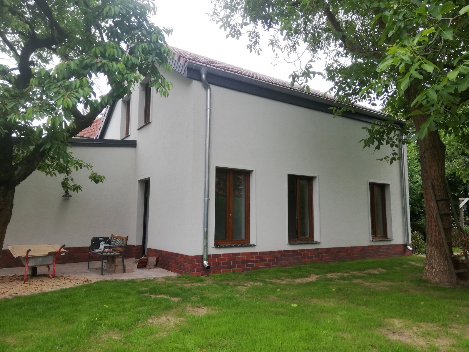 Świeżo otynkowany dom z ceglaną podmurówką, drewnianymi oknami i metalowymi rynnami, widok z zewnątrz na trawnik i drzewa.