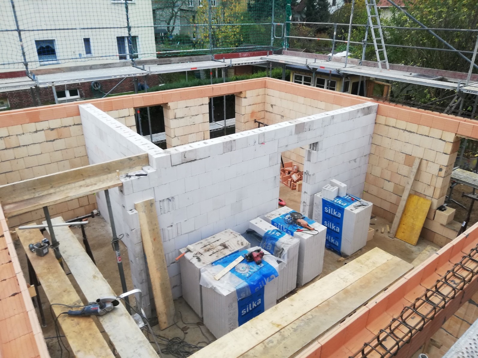 Wznoszenie ścian parteru domu z bloczków silikatowych i ceramiki poryzowanej, widoczne narzędzia i materiały budowlane na placu budowy, rusztowania w tle.