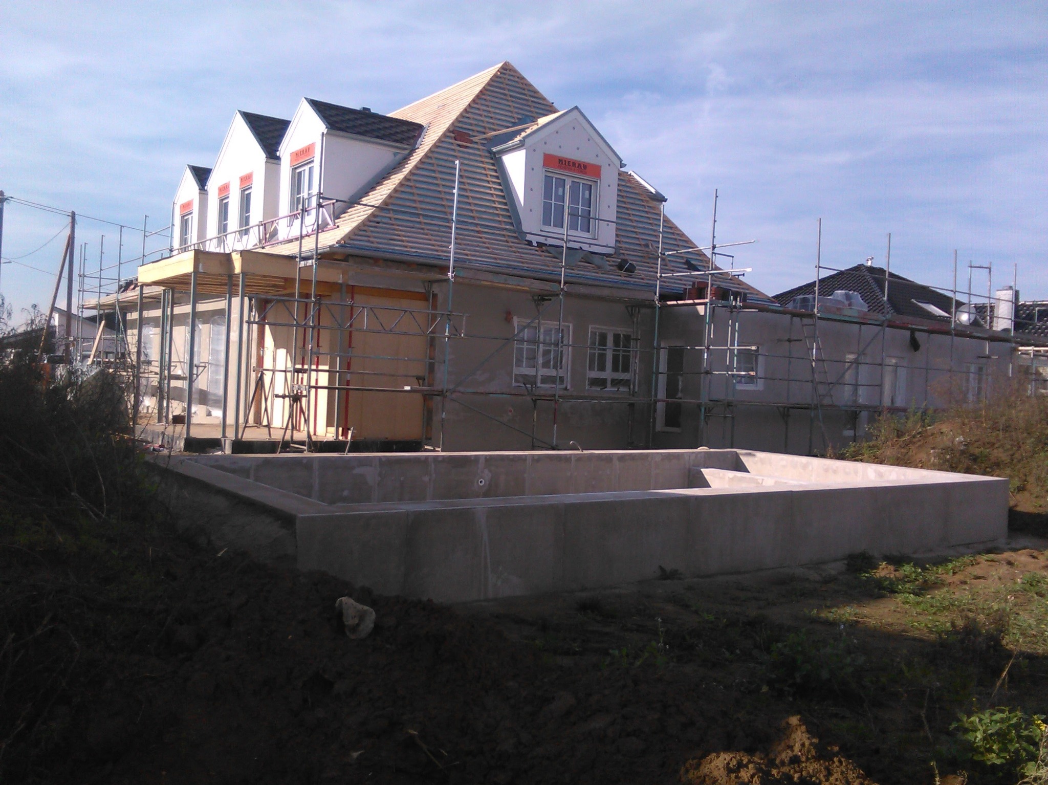 Budowa domu w trakcie realizacji: wylany fundament, ściany w stanie surowym, częściowo położona dachówka, rusztowania wokół budynku.