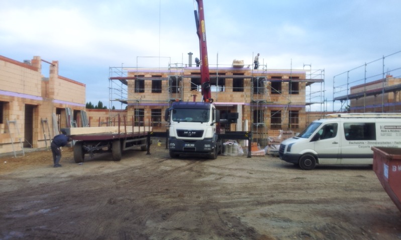 Wznoszenie dwukondygnacyjnego budynku z pustaków ceramicznych, z użyciem dźwigu samochodowego i rusztowań, widoczny pracownik przyczepy i samochód dostawczy z reklamą firmy budowlanej.