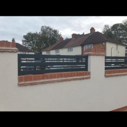 Sebastian Kaca - Nowoczesne ogrodzenie z poziomych, grafitowych elementów metalowych, osadzone na słupkach z białego tynku i cegły klinkierowej, na tle domów jednorodzinnych.