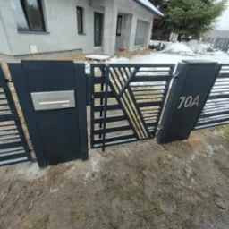 Nowoczesna, czarna brama wjazdowa z geometrycznym wzorem, numerem domu 70A i skrzynką na listy, na tle surowego stanu budynku i resztek śniegu.