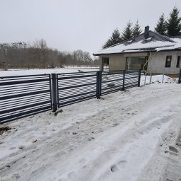 Sebastian Kaca - Nowoczesne ogrodzenie panelowe z poziomymi szczeblami w kolorze grafitowym, otoczone śnieżnym krajobrazem przed domem w trakcie budowy.