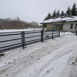 Nowoczesne ogrodzenie panelowe z poziomymi szczeblami w kolorze grafitowym, otoczone śnieżnym krajobrazem przed domem w trakcie budowy.