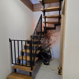 Sebastian Kaca - Drewniane schody z czarną metalową konstrukcją i barierką, z zabezpieczeniem dla dzieci na dole. W tle tapeta z motywem dmuchawca i wózek dziecięcy.