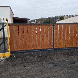 Sebastian Kaca - Nowoczesna brama przesuwna z drewnopodobnymi panelami pionowymi i szarą ramą, z żółtymi elementami automatyki, na tle domu z ogrodzeniem panelowym.