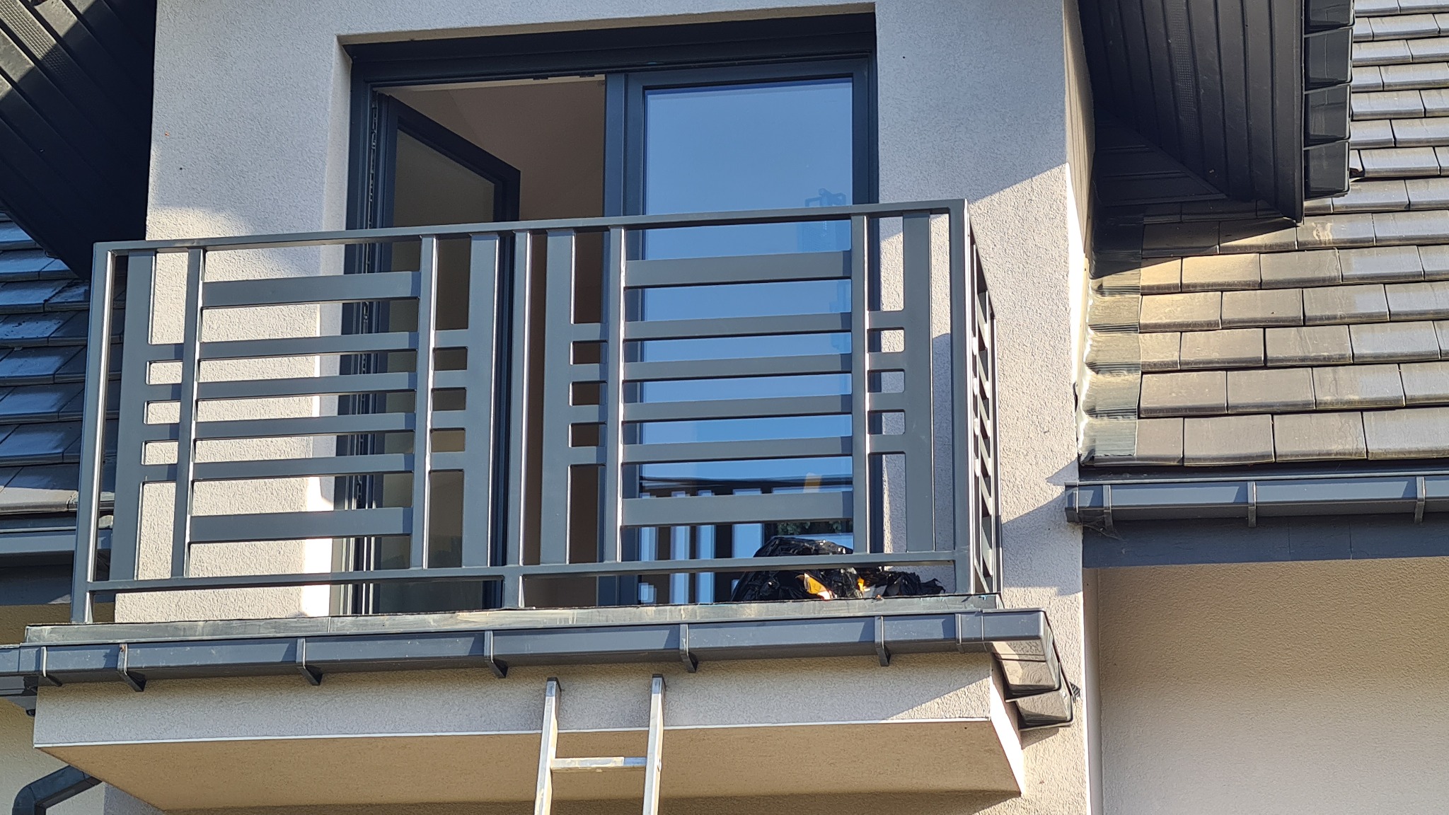 Nowoczesny balkon z szarą, metalową balustradą o geometrycznym wzorze, widok z dołu na tle jasnej elewacji i fragmentu dachu z dachówką ceramiczną, drabina oparta o ścianę pod balkonem.