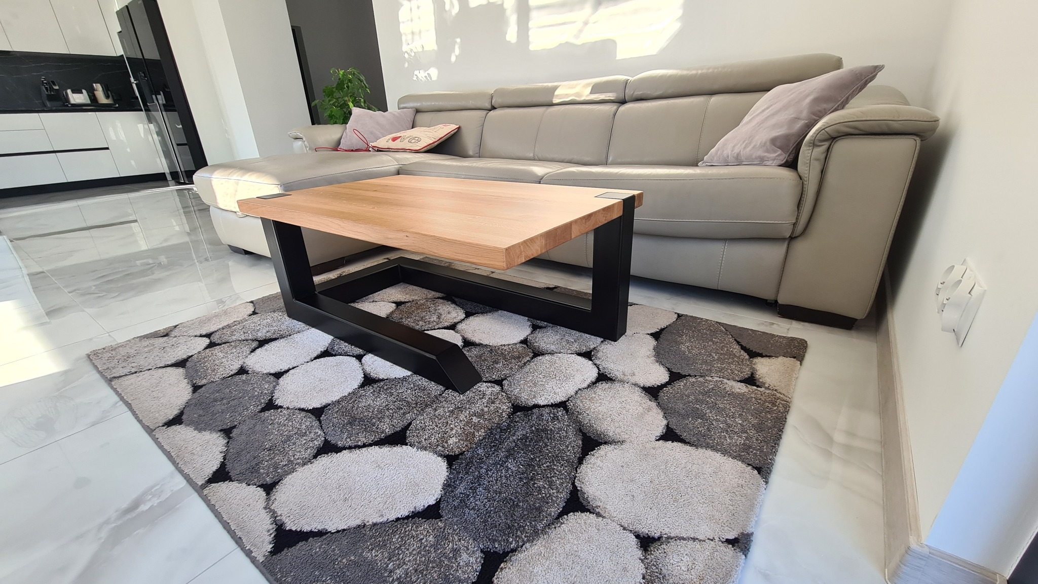 Stolik kawowy z drewnianym blatem i czarną, metalową podstawą, umieszczony na dywanie imitującym kamienie, w tle beżowa sofa w nowoczesnym salonie.