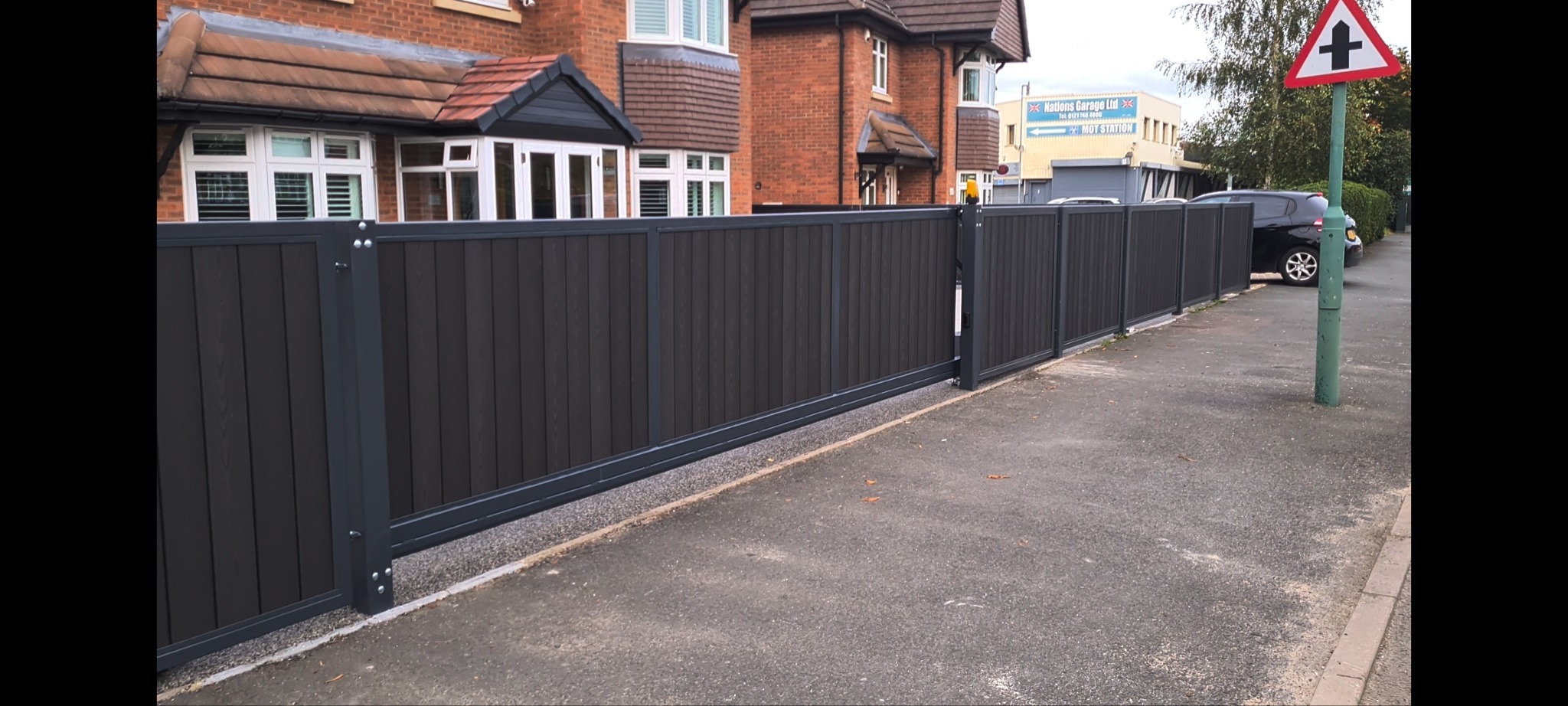 Nowoczesna, przesuwna brama wjazdowa z ciemnego drewna kompozytowego i szarej metalowej ramy, widoczna z chodnika na tle domu z czerwonej cegły i budynku z szyldem reklamowym.